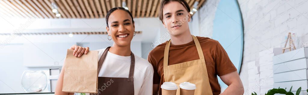 Smiling african american seller holding paper bag near colleague with coffee to go in sweet shop, banner 