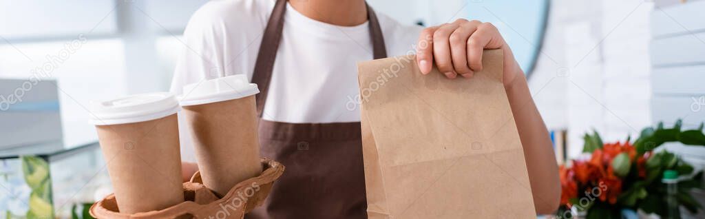 Cropped view of african american worker holding coffee to go and paper bag in sweet shop, banner 