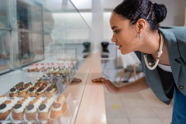 Side view of smiling african american client choosing eclairs near showcase in sweet shop 
