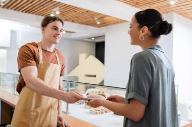 Positive seller giving dessert to african american client in sweet shop 