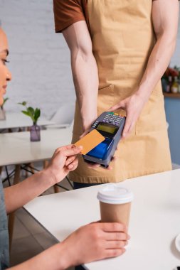 Salesman holding payment terminal near african american client with coffee in sweet shop 