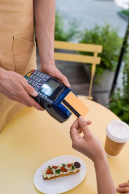 Cropped view of african american woman paying with credit card near seller and dessert on terrace of sweet shop 