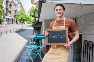 Smiling salesman holding chalkboard with franchise lettering near confectionery outdoors 