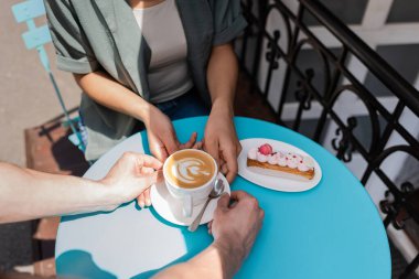 Cropped view of seller putting coffee on table near eclair and african american client on terrace of cafe 