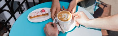 Cropped view of salesman putting cappuccino near eclair and african american woman on terrace of sweet shop, banner 