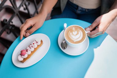 Top view of african american woman holding cappuccino near eclair on terrace of sweet shop 