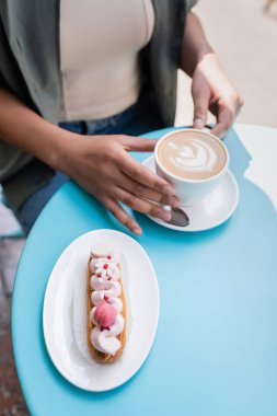 High angle view of delicious eclair near blurred african american woman with cup of cappuccino on terrace of confectionery 