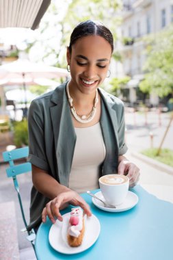 Gülümseyen Afrikalı Amerikalı kadın elinde cappuccino ile şekerci dükkanının terasında eclair alıyor. 