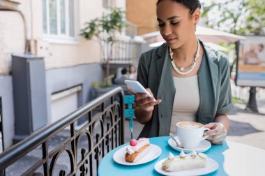 African american woman using smartphone near coffee and eclairs on terrace of sweet shop 