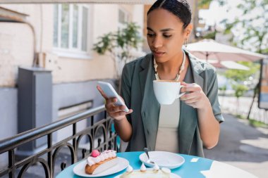 African american woman using smartphone and holding cappuccino near eclairs on terrace of sweet shop 