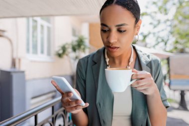 Genç Afrikalı Amerikalı kadın elinde bir fincan kapuçino tutuyor ve şekerci dükkanının terasında akıllı telefon kullanıyor. 