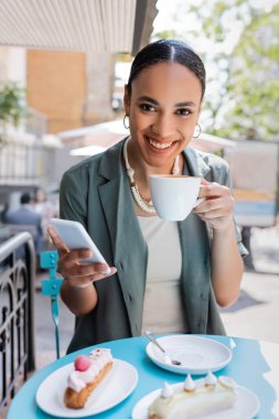 Smiling african american client holding coffee and smartphone near eclairs on terrace of sweet shop 