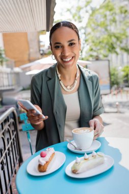 Pretty african american client holding smartphone near cappuccino and eclairs on terrace of confectionery 