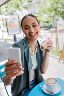 Smiling african american woman holding eclair and taking selfie on smartphone near cappuccino on terrace of sweet shop 