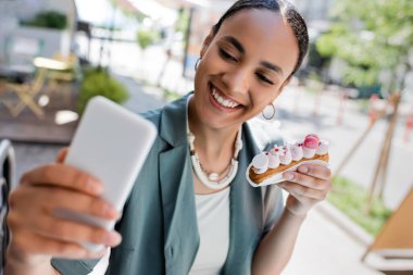 Cheerful african american woman holding eclair and taking selfie on smartphone on terrace of confectionery 