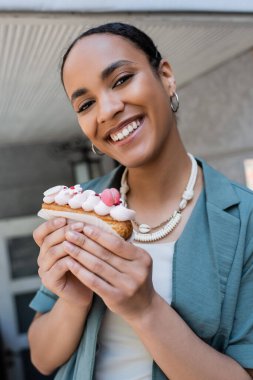 Cheerful african american woman holding delicious eclair and looking at camera outdoors  clipart