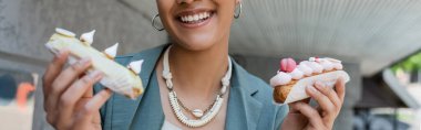 Cropped view of smiling african american client holding eclairs outdoors, banner 