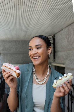 Neşeli genç Afrikalı Amerikalı kadın şekerci dükkanının yanında kremalı ekler tutuyor. 