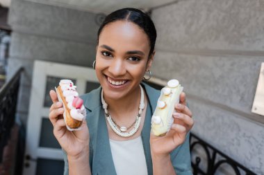 African american customer holding eclairs and looking at camera on terrace of sweet shop 