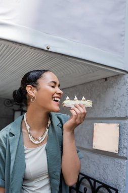 Gülümseyen Afro-Amerikan kadın, dışarıda şekerci dükkanının yanında eclair tutarken gözlerini kapatıyor. 