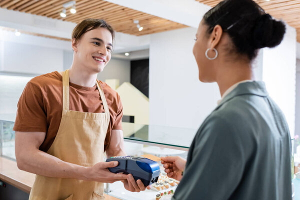 Cheerful seller holding payment terminal near african american client with credit card in sweet shop 