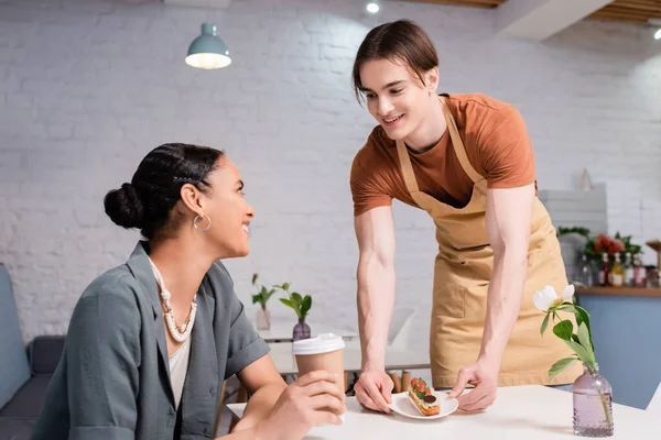 Cheerful seller giving dessert to african american customer with coffee to go in confectionery