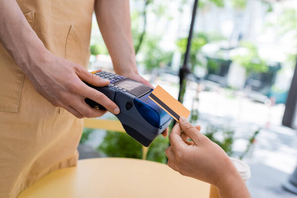 Cropped view of african american client paying with credit card near seller and paper cup on terrace of confectionery 
