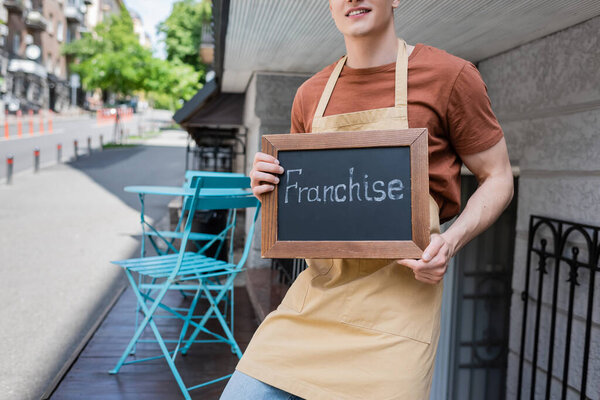 Cropped view of smiling seller holding chalkboard with franchise lettering near sweet shop on urban street 