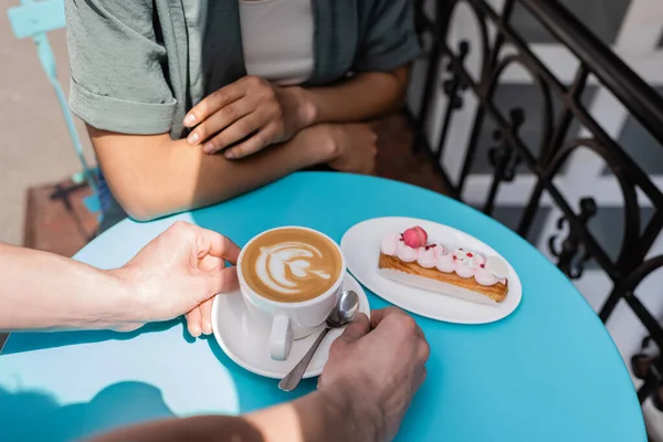 Cropped view of seller putting cappuccino on table near dessert and african american client on terrace of sweet shop  