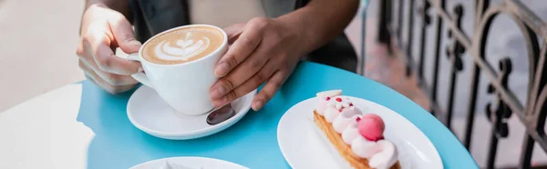 Pemandangan Dari Wanita Afrika Amerika Memegang Cangkir Cappuccino Dekat Eclair — Stok Foto