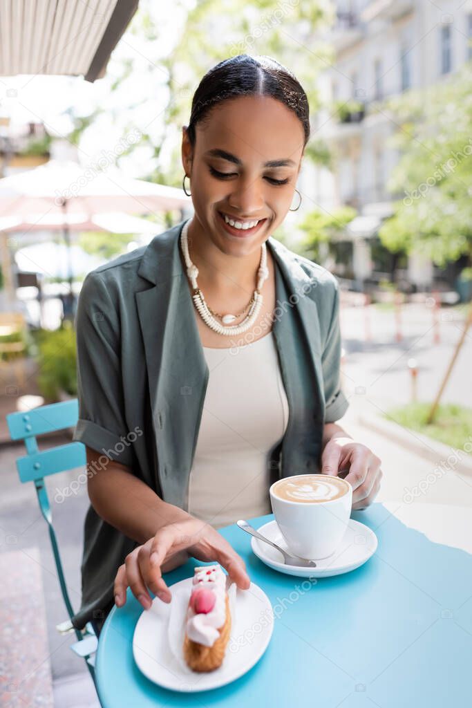 Smiling african american woman holding cappuccino and taking eclair on terrace of sweet shop 