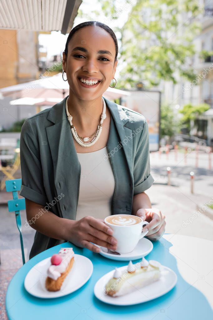 Smiling african american woman looking at camera near cappuccino and eclairs on terrace of confectionery 
