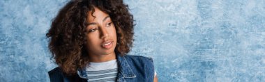 smiling african american woman in denim vest looking away on blue background, banner