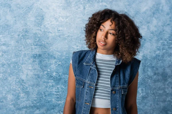 african american woman in denim vest and striped crop top looking away on blue textured background