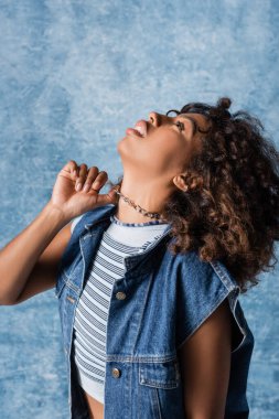 african american woman in denim vest touching necklace and looking up on blue background