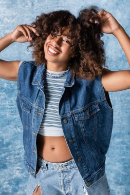 happy african american woman in denim vest touching wavy hair and looking at camera on blue background