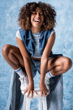 laughing african american woman in denim clothing posing on haunches on blue background