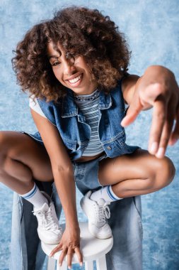 happy african american woman in denim clothes pointing at camera while sitting on haunches on blue background