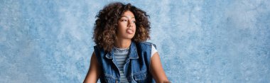 curly african american woman in denim vest looking away on blue background, banner