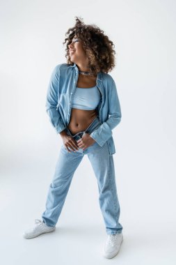 full length of african american woman in denim clothes posing with thumbs in belt loops on grey background