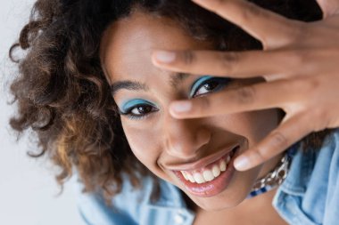 cheerful african american woman with makeup obscuring face with hand isolated on blue