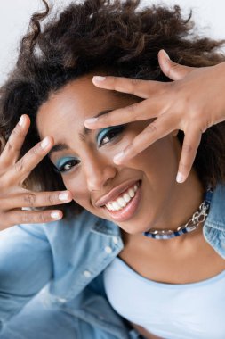 happy african american woman obscuring face with hands while looking at camera isolated on grey