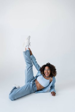Heyecanlı, mavi kot pantolon giymiş, gri arka planda bacağı kalkık, Afro-Amerikan kadın.