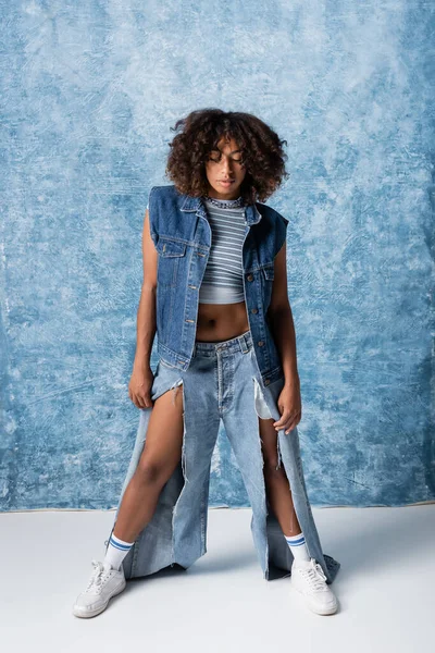full length of african american woman in torn jeans and white sneakers standing on blue background