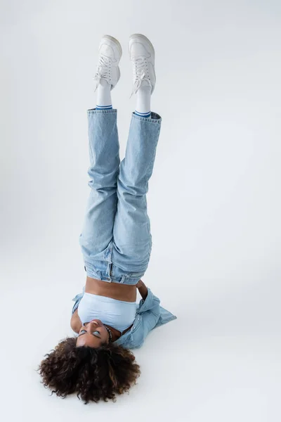 african american woman in blue jeans and white sneakers in shoulder stand pose on grey background