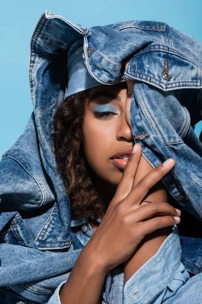 african american woman with makeup obscuring face with denim jacket isolated on blue