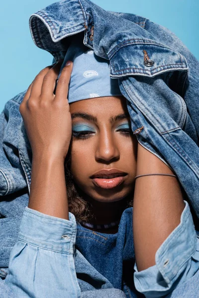 african american woman with makeup covering head with denim jacket isolated on blue