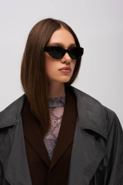 brunette teenage model in stylish sunglasses and trendy trench coat isolated on grey
