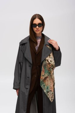 brunette teenage girl in stylish sunglasses and trendy trench coat isolated on grey