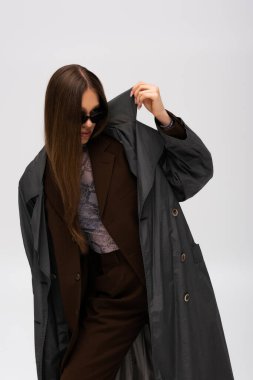 stylish teenage model in sunglasses adjusting collar of trendy trench coat isolated on grey